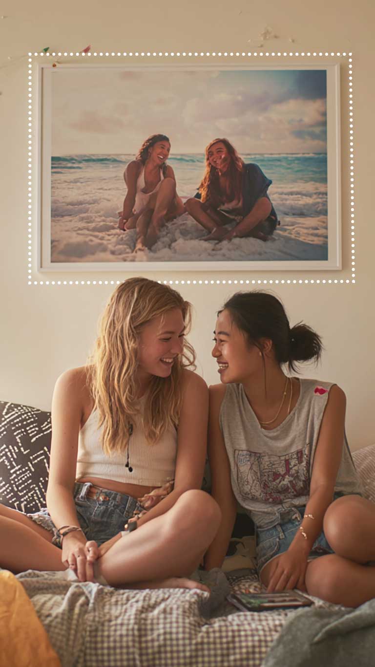 Two young women smile at each other on a bed beneath a large poster of themselves together on a beach