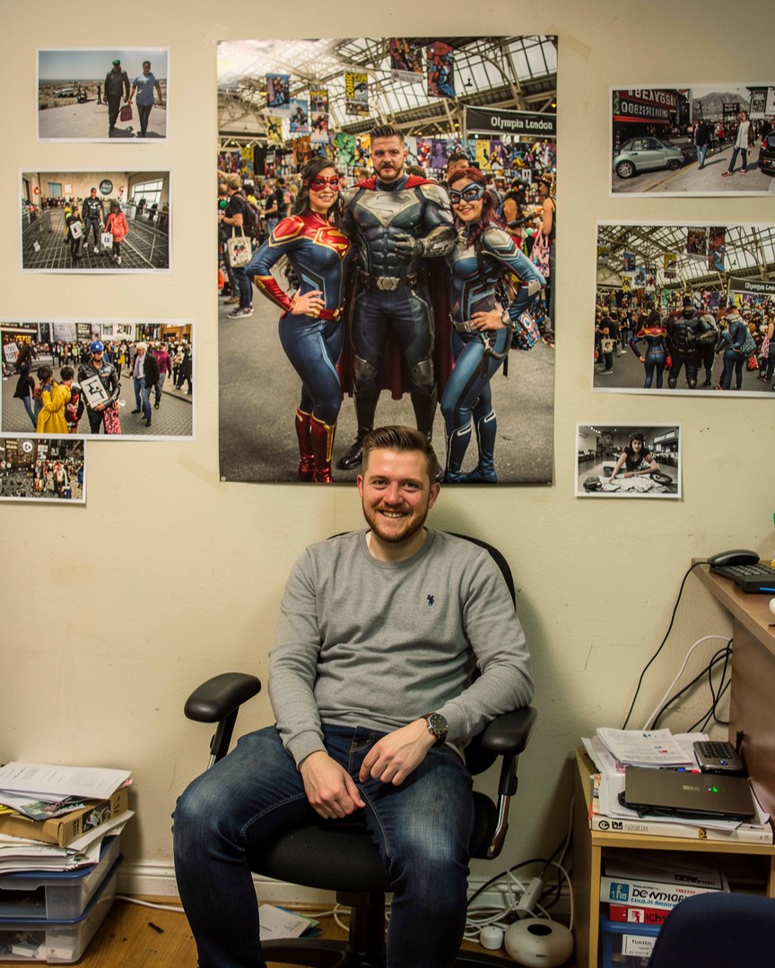 A smiling man in an office setting sits in front of a large wall poster featuring three people in superhero cosplay at a Comic-Con event.