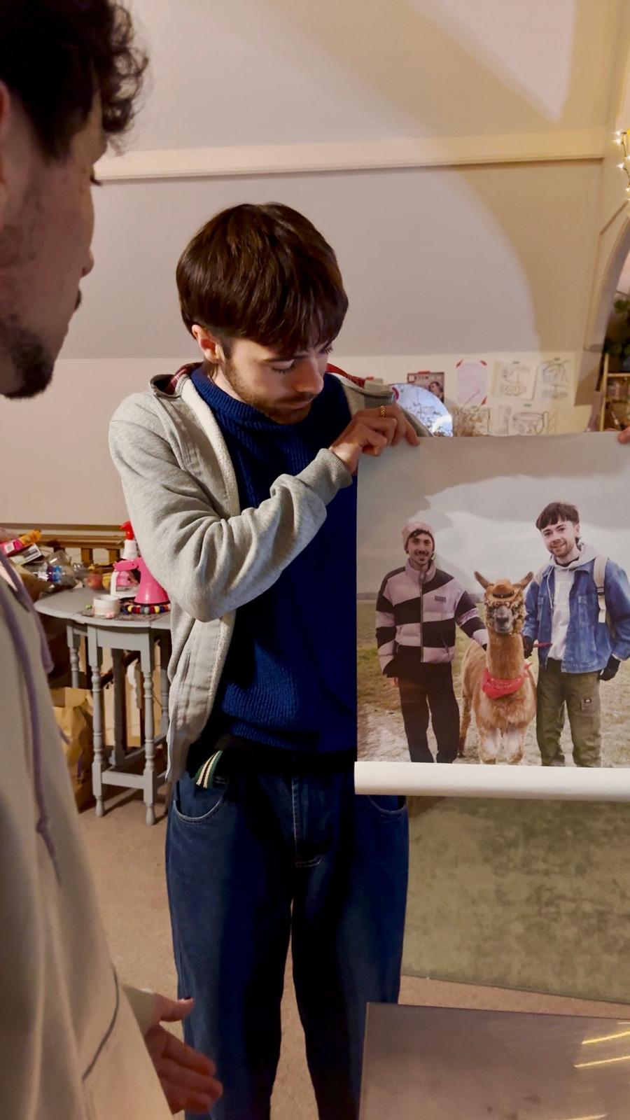 A young man holds up a poster print showing himself and a friend standing in a field with a llama wearing a small hat and a red bandana.